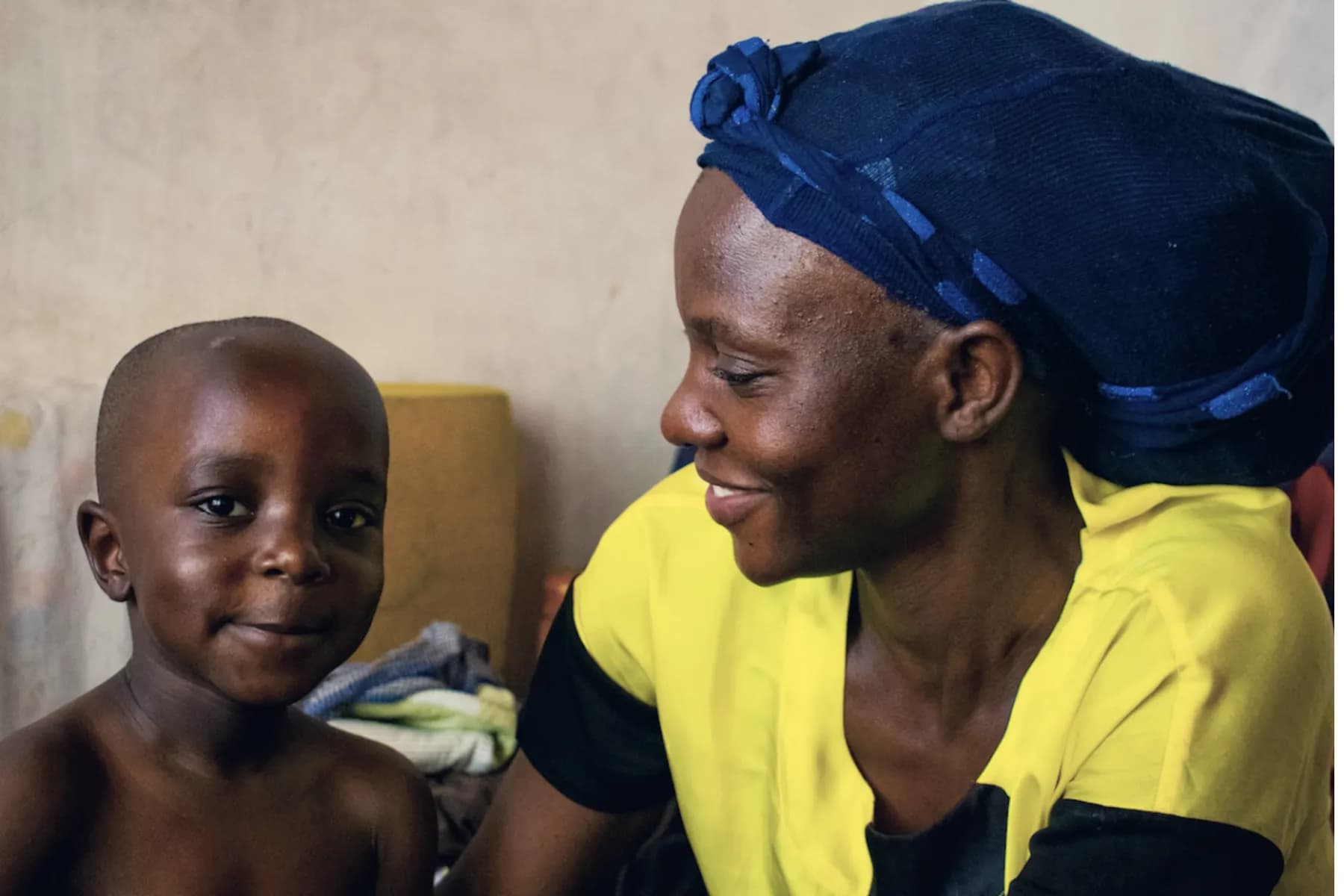 A mother in a blue headwrap smiling beside her young son